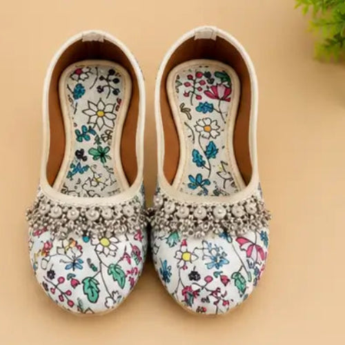 A pair of women's flat shoes with a white base, colorful floral print, and a silver rhinestone embellished band across the vamp, displayed on a beige background with a green plant in the corner.