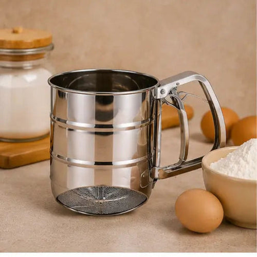 A stainless steel flour sifter with a handle and mesh bottom, shown on a beige countertop with eggs, a bowl of flour, and a jar of sugar in the background.