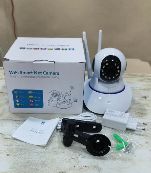 White wireless smart security camera with two antennas, shown next to its white retail box, a black mounting bracket, a white wall adapter, a power cord, and a small instruction booklet on a light-colored wooden surface.